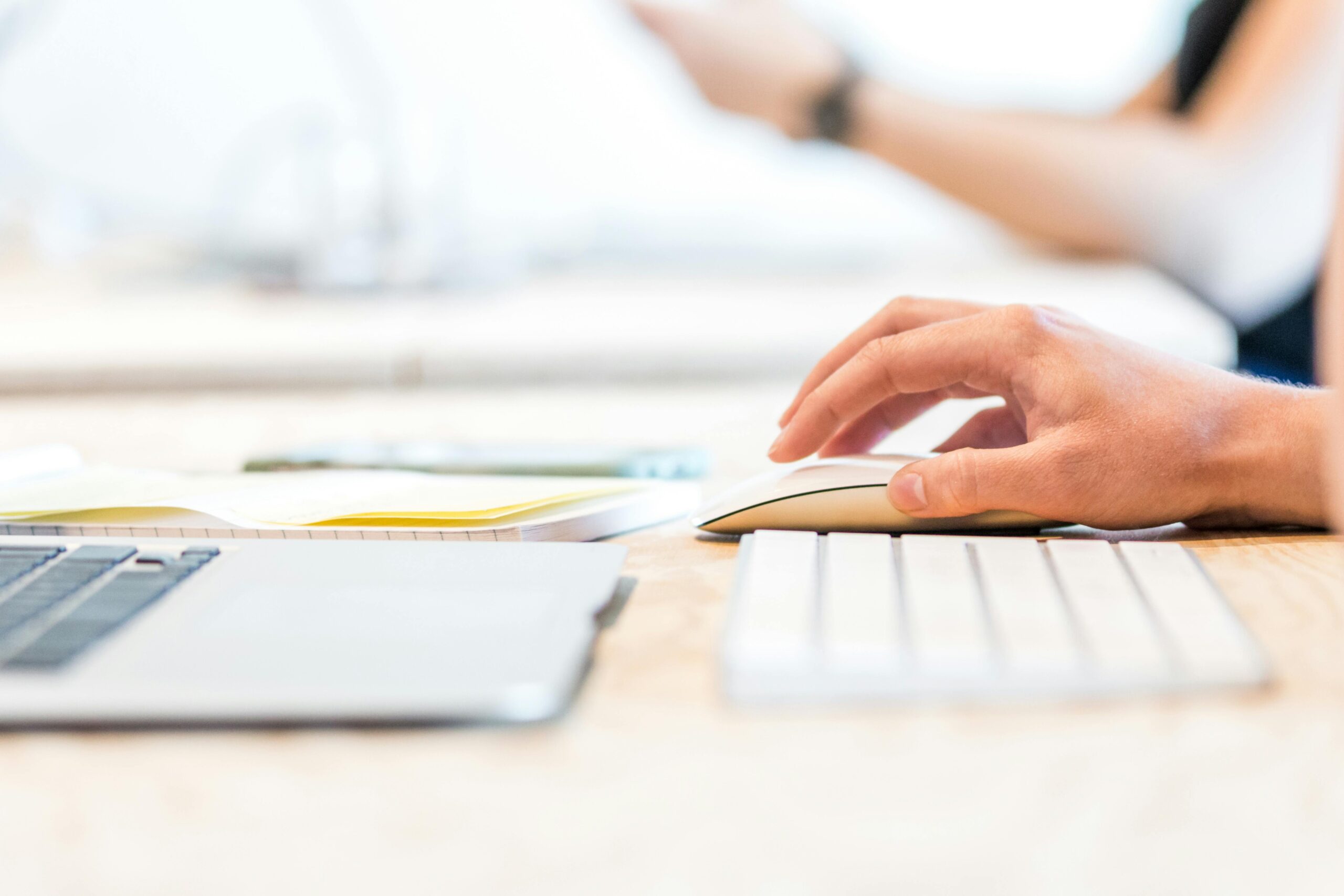 Close-up of a hand using a computer mouse next to a laptop and keyboard in a bright, minimalist workspace.